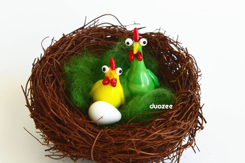 Peut inclure: Un nid tiss&eacute; brun avec deux poulets en verre, un jaune et un vert, et un &oelig;uf blanc. Les poulets ont de grands yeux globuleux.