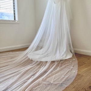 K&ouml;nnte beinhalten: Ein langer, transparenter wei&szlig;er Brautschleier aus feinem T&uuml;llstoff mit einem zarten Muster aus kleinen, gleichm&auml;&szlig;ig verteilten wei&szlig;en Schleifen entlang des geschwungenen Saums, der elegant von oben bis zum Boden herabf&auml;llt.