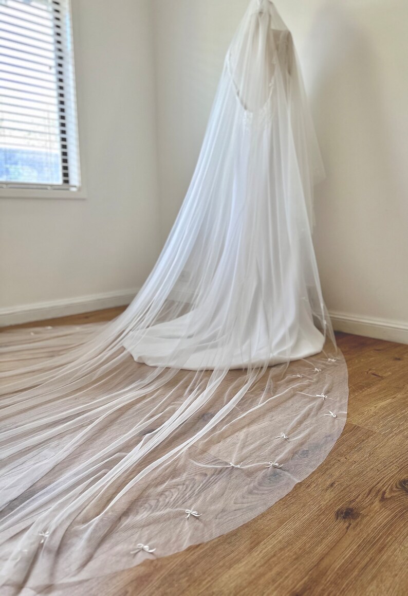 K&ouml;nnte beinhalten: Ein langer, transparenter wei&szlig;er Brautschleier aus feinem T&uuml;llstoff mit einem zarten Muster aus kleinen, gleichm&auml;&szlig;ig verteilten wei&szlig;en Schleifen entlang des geschwungenen Saums, der elegant von oben bis zum Boden herabf&auml;llt.