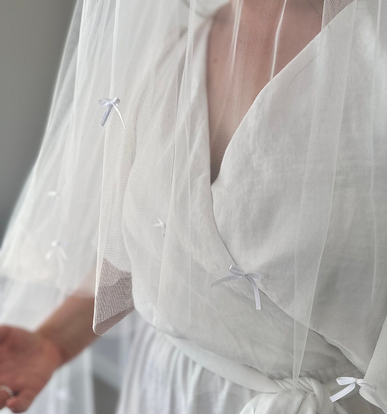 K&ouml;nnte beinhalten: Ein wei&szlig;es Wickelkleid mit V-Ausschnitt und Bindeg&uuml;rtel, kombiniert mit einem transparenten wei&szlig;en T&uuml;llschleier. Der Schleier ist mit kleinen, gleichm&auml;&szlig;ig verteilten wei&szlig;en Schleifen aus Band verziert, was f&uuml;r ein zartes Aussehen sorgt.