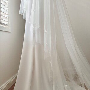 K&ouml;nnte beinhalten: Ein bodenlanges wei&szlig;es Kleid mit einer glatten, blickdichten Stoffbasis und einem transparenten, mehrlagigen T&uuml;llschleier. Der T&uuml;ll&uuml;berwurf ist mit kleinen, zarten wei&szlig;en Schleifen aus Band verziert, die &uuml;ber den gesamten Stoff verteilt sind.