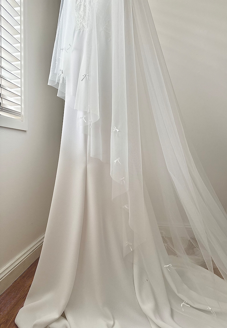 K&ouml;nnte beinhalten: Ein bodenlanges wei&szlig;es Kleid mit einer glatten, blickdichten Stoffbasis und einem transparenten, mehrlagigen T&uuml;llschleier. Der T&uuml;ll&uuml;berwurf ist mit kleinen, zarten wei&szlig;en Schleifen aus Band verziert, die &uuml;ber den gesamten Stoff verteilt sind.