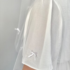 K&ouml;nnte beinhalten: Ein wei&szlig;es, transparentes Kleidungsst&uuml;ck mit kurzen, lockeren &Auml;rmeln, die mit zarten, kleinen wei&szlig;en Schleifen aus Band verziert sind. Das Material ist leicht und halbtransparent, mit einer weichen Textur und einer klaren, hellwei&szlig;en Farbe.