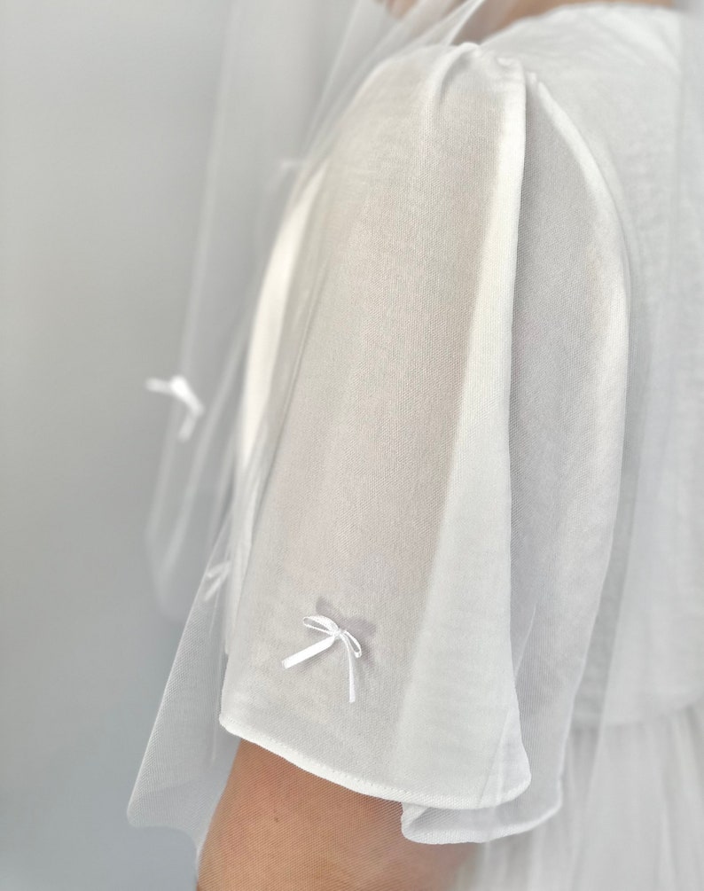 K&ouml;nnte beinhalten: Ein wei&szlig;es, transparentes Kleidungsst&uuml;ck mit kurzen, lockeren &Auml;rmeln, die mit zarten, kleinen wei&szlig;en Schleifen aus Band verziert sind. Das Material ist leicht und halbtransparent, mit einer weichen Textur und einer klaren, hellwei&szlig;en Farbe.