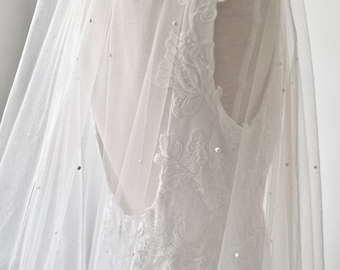 Könnte beinhalten: Ein weißer Hochzeitsschleier aus transparentem Tüllstoff mit über das Material verteilten kleinen weißen Perlen. Der Schleier liegt über einem ärmellosen weißen Kleid, das an der Seite und am Rücken mit aufwendigen weißen Blumenstickereien versehen ist.