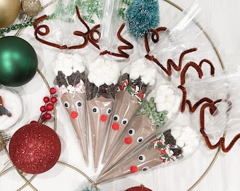 May include: Five cone-shaped bags filled with hot chocolate mix, marshmallows, and toppings, decorated to resemble reindeer. Each bag has googly eyes and a red nose. Brown pipe cleaner antlers and Christmas ornaments complete the festive scene.
