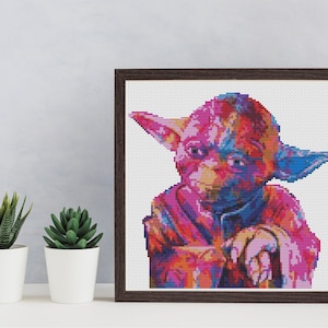 Peut inclure: Un point de croix encadré représentant un portrait abstrait et coloré de Yoda de Star Wars. Le fond est blanc et le cadre est brun.