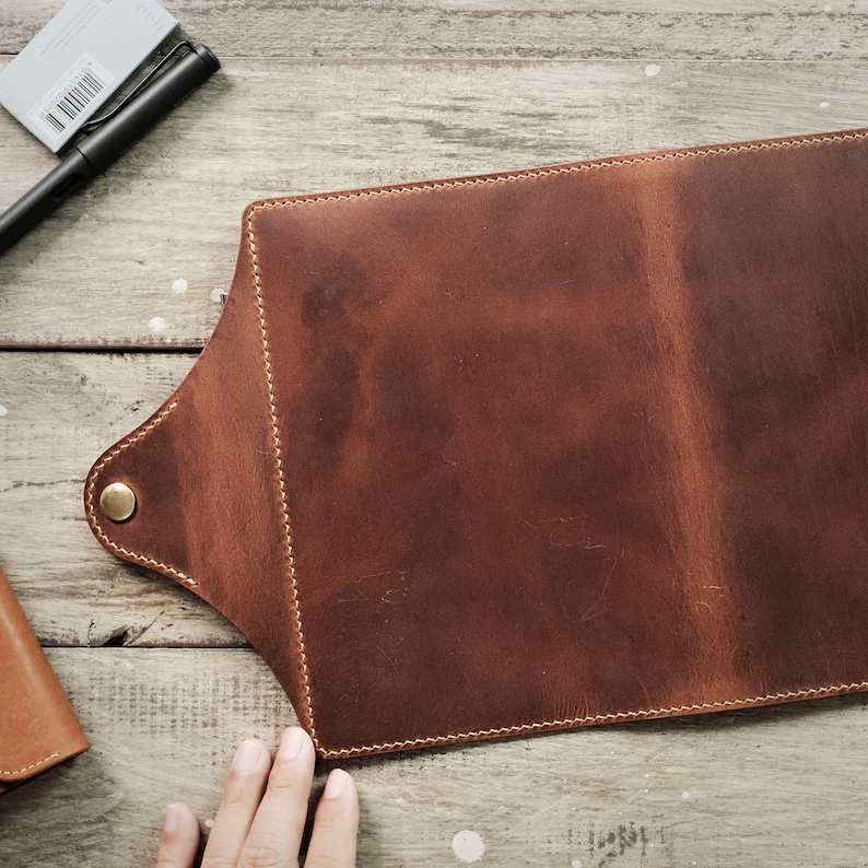 May include: A brown leather notebook cover with a flap and a snap closure. The cover has a distressed look and is stitched with a light brown thread.