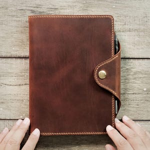 May include: A brown leather notebook with a flap closure and a brass button. The notebook is closed and has a pen tucked into the flap.