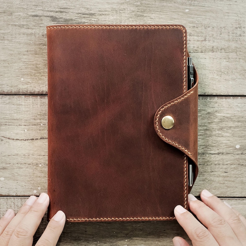 May include: A brown leather notebook with a flap closure and a brass button. The notebook is closed and has a pen tucked into the flap.