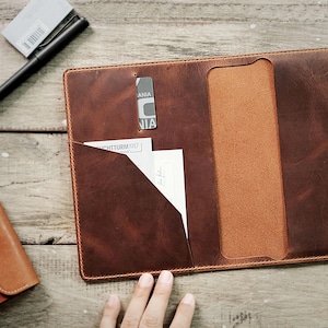 May include: A brown leather portfolio with a flap closure and a pocket on the front. The portfolio is open and a piece of paper is visible in the pocket. The portfolio is on a wooden surface.
