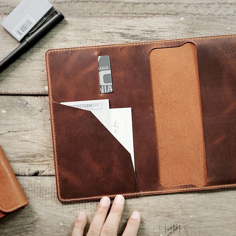 May include: A brown leather portfolio with a flap closure and a pocket on the front. The portfolio is open and a piece of paper is visible in the pocket. The portfolio is on a wooden surface.