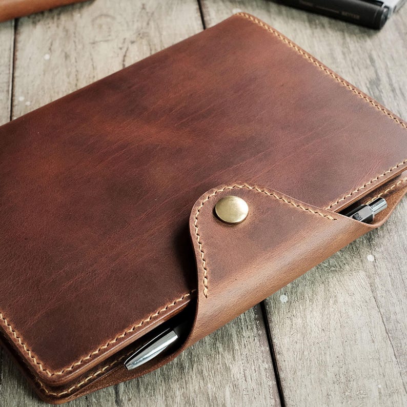 May include: A brown leather notebook cover with a pen holder and a brass button closure.