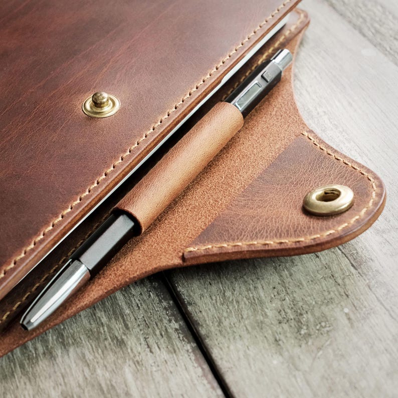 May include: A brown leather notebook cover with a pen tucked into the flap. The cover has a stitched edge and a brass button closure.