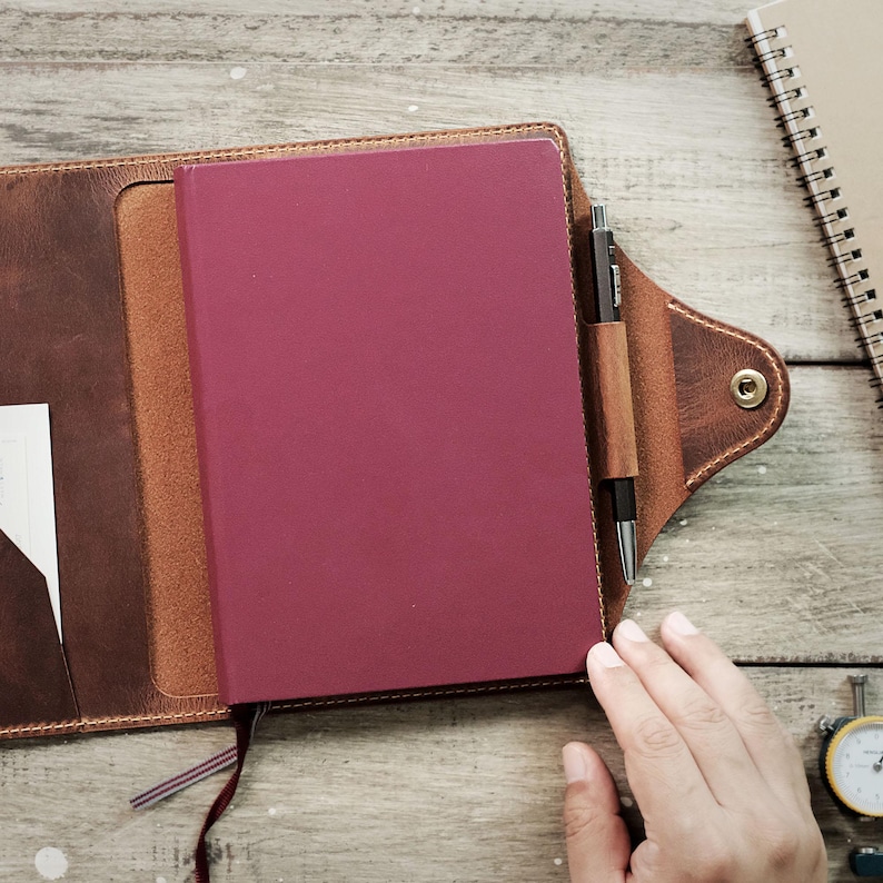 May include: A brown leather notebook cover with a burgundy notebook inside. A pen is tucked into a leather pocket on the cover.