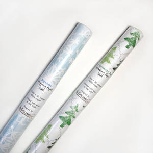 May include: Two rolls of white wrapping paper with different designs. One roll features a snowflake pattern in light blue and the other features a green Christmas tree pattern. Both rolls are 1 foot by 30 inches and are made of matte paper. The text on the rolls reads "Snowflake Wrapping Paper Roll" and "Christmas Tree Wrapping Paper Roll". Both rolls are printed with love in the USA and designed by Schmidt.