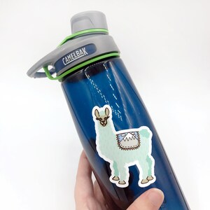 Puede incluir: Una botella de agua azul con una tapa verde y una pegatina de una llama azul claro con un dise&ntilde;o de monta&ntilde;a en la espalda. La pegatina est&aacute; en el lateral de la botella. La botella tiene la palabra "CamelBak" en ella.