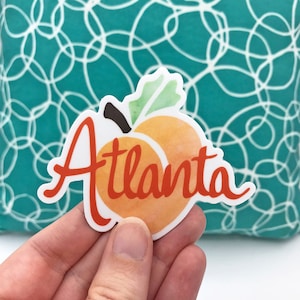 May include: A peach-colored sticker with the word "Atlanta" in red lettering. The peach has a green leaf and a brown stem.