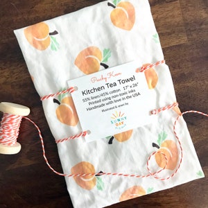 May include: A white kitchen tea towel with a peach design. The towel is 17 inches by 26 inches and is made of 55% linen and 45% cotton. The towel is tied with twine and has a tag that says "Peachy Keen Kitchen Tea Towel" and "Sunny Day Designs".