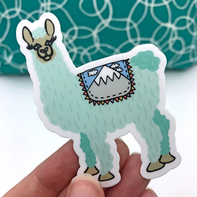 Puede incluir: Una pegatina de llama azul claro con un dise&ntilde;o de monta&ntilde;a colorido en su espalda. La llama tiene una cara blanca con pesta&ntilde;as negras y una nariz rosa.