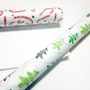 May include: Two rolls of white wrapping paper with different Christmas designs. One roll has red candy canes and the other has green Christmas trees.