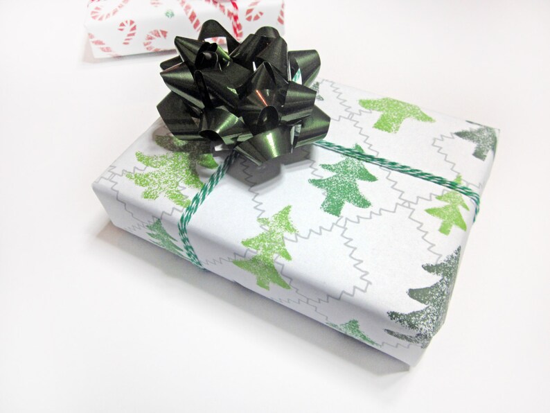 May include: A white gift box wrapped in green Christmas tree patterned paper with a black satin bow. The box is tied with green twine.