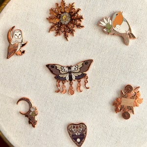 May include: Set of ten enamel pins featuring various designs, including a sun, a moth, a bird, a crescent moon, a heart, and a spool of thread. The pins are all in a copper tone.