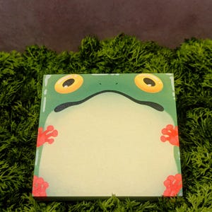 May include: Square notepad with a green frog face design. The frog has large yellow eyes and red feet. The notepad is set against a background of green moss.