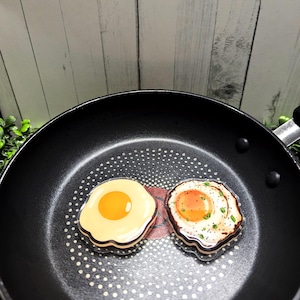 Puede incluir: Dos objetos decorativos con forma de huevo frito en una sartén negra. Un huevo tiene una yema amarilla, el otro tiene una yema cocida con pimienta. La sartén tiene una superficie antiadherente con puntos blancos y un centro rojo.