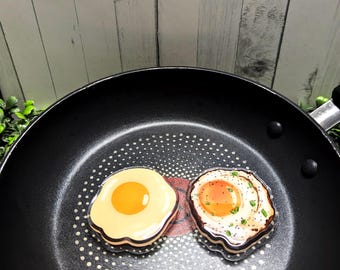May include: Two fried egg-shaped decorative items in a black frying pan. One egg has a yellow yolk, the other has a cooked yolk with pepper. The pan has a non-stick surface with white dots and a red center.