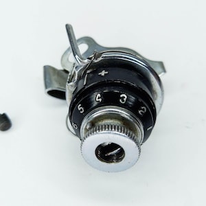 May include: A sewing machine tension assembly with a black dial marked with white numbers from 2 to 5. The assembly is silver and black, with a small screw to the left. The background is white.