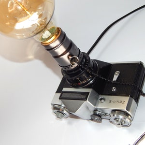 Könnte beinhalten: Eine einzigartige Schreibtischlampe aus einer Vintage Zenith-E Kamera. Die Lampe verfügt über eine große Edison-Glühbirne und ein schwarzes Netzkabel. Das Kameragehäuse ist silber und schwarz, mit dem Text "ZENIT-E".