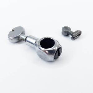 May include: Two small, metallic machine parts on a white background. One part is a silver-colored, cylindrical piece with a circular opening and a slotted head. The other is a smaller, hook-shaped piece, also metallic.