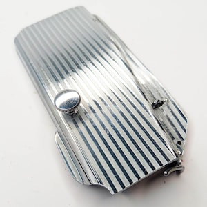 May include: A silver-colored, striped, folding pocket knife with a small round knob on the top. The knife is closed and has a hinged clasp.