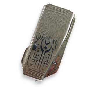 May include: A silver-toned, rectangular cigarette case with intricate engraved patterns. The design features geometric shapes and stylized floral motifs. A small, round button is visible on the front, and a hinged lid is slightly ajar.
