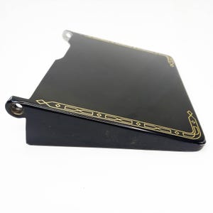 May include: A black, angled metal piece with a glossy finish. It features a gold geometric design along the edges. The piece has a rectangular shape with a cut-out section and two circular holes.