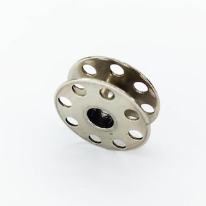 May include: A silver metal bobbin with a circular shape and multiple holes around the edge. The bobbin is designed for use in sewing machines.