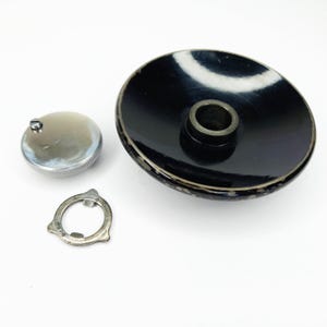 May include: A collection of vintage metal parts, including a large black circular piece with a central cylinder, a small silver cap, and a silver ring with the text "#5716". The parts are arranged on a white surface.