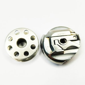 May include: A silver sewing machine bobbin case and a silver bobbin with a circular design and eight holes. The bobbin case has the text "SINGER 48780" printed on it.