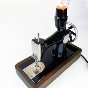 Può includere: Una lampada vintage a forma di macchina da cucire nera con base in legno. La lampada è accesa e la lampadina brilla.