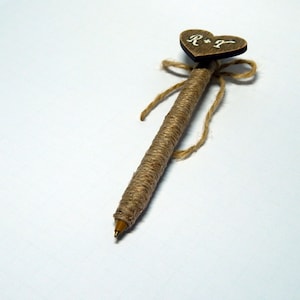 Może przedstawiać: Brązowe drewniane serce z napisem "R+J" w kolorze białym przymocowane do długopisu owiniętego sznurkiem. Długopis jest związany kokardką ze sznurka.