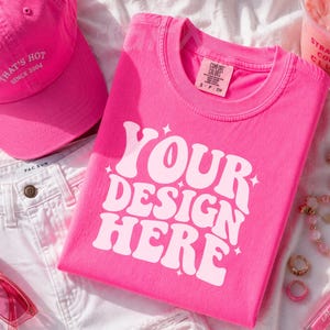 May include: A vibrant pink t-shirt with the text "YOUR DESIGN HERE" is folded on a white surface. A matching pink baseball cap, sunglasses, and accessories like jewelry and a drink are also present, creating a coordinated aesthetic.