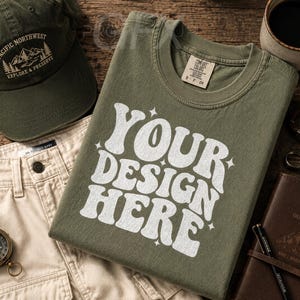 May include: An olive green t-shirt with the text "YOUR DESIGN HERE" in white, alongside a matching cap, khaki pants, a compass, and a coffee mug. A leather journal and accessories complete the outdoor theme.