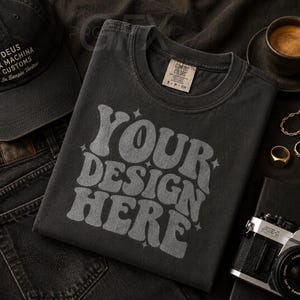 May include: A black t-shirt with the text "YOUR DESIGN HERE" in a retro font, a black baseball cap, and a pair of black jeans. The image also includes a cup of coffee, gold jewelry, and a vintage camera, all arranged on a dark surface.