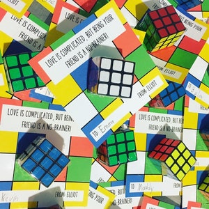 Könnte beinhalten: Eine Sammlung bunter Karten mit dem Text "Liebe ist kompliziert, aber dein Freund zu sein ist ein Kinderspiel!" Jede Karte hat einen kleinen, bunten Rubik's Cube daran befestigt. Die Karten liegen auf einem karierten Hintergrund.