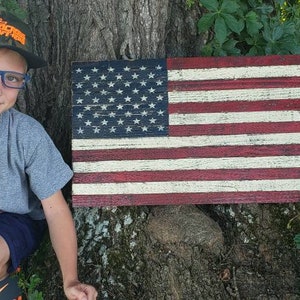 May include: A young person wearing a camouflage baseball cap with an orange logo and blue-rimmed glasses sits in front of a wooden American flag. The flag is painted in red, white, and blue with a distressed look. The person is wearing a grey t-shirt with the words "Go Off Road" printed on it.