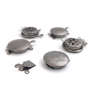 Puede incluir: Seis cierres de metal plateados para la elaboración de joyas. Los cierres son redondos con un diseño decorativo y tienen un bucle a cada lado. Un cierre es un clip con un bucle a un lado.