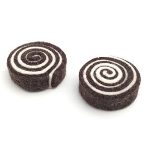 May include: Two felted wool coasters. Each coaster is a dark brown cylinder with a white spiral design on top. The coasters are on a white background.