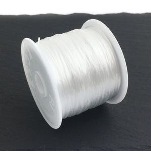 May include: A spool of clear, elastic beading cord. The cord is tightly wound on a white plastic spool. The spool is resting on a dark, textured surface. This cord is ideal for jewelry making and crafting projects.