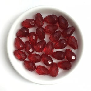 以下が含まれることがあります： 深い赤色の、面カットされたしずく型のビーズのコレクション。白い浅いボウルに入っています。ビーズは光沢があり、ボウルを満たすように配置されており、視覚的に魅力的なディスプレイになっています。ジュエリー作りに最適です。